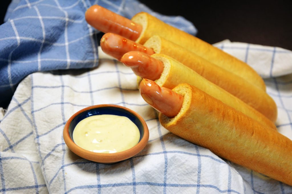 Cztery gotowe hot dogi francuskie z parówką w dziurce leżą na niebiesko-białej ściereczce obok miseczki z jasnym sosem