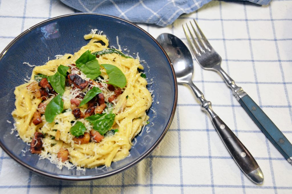 Makaron z boczkiem, szpinakiem i startym parmezanem w niebieskiej misce na kraciastej ściereczce