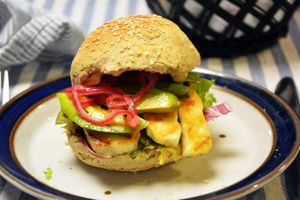 Burger w bułce sezamowej z grillowanymi plastrami halloumi, awokado, marynowaną czerwoną cebulą i sałatą na talerzu