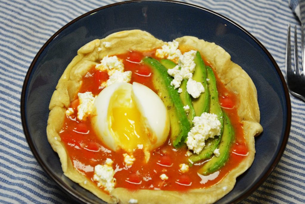 Huevos rancheros w głębokiej niebieskiej misce: miękka tortilla pszenna z salsą roja, przekrojone jajko na półtwardo, plastry awokado i pokruszona feta
