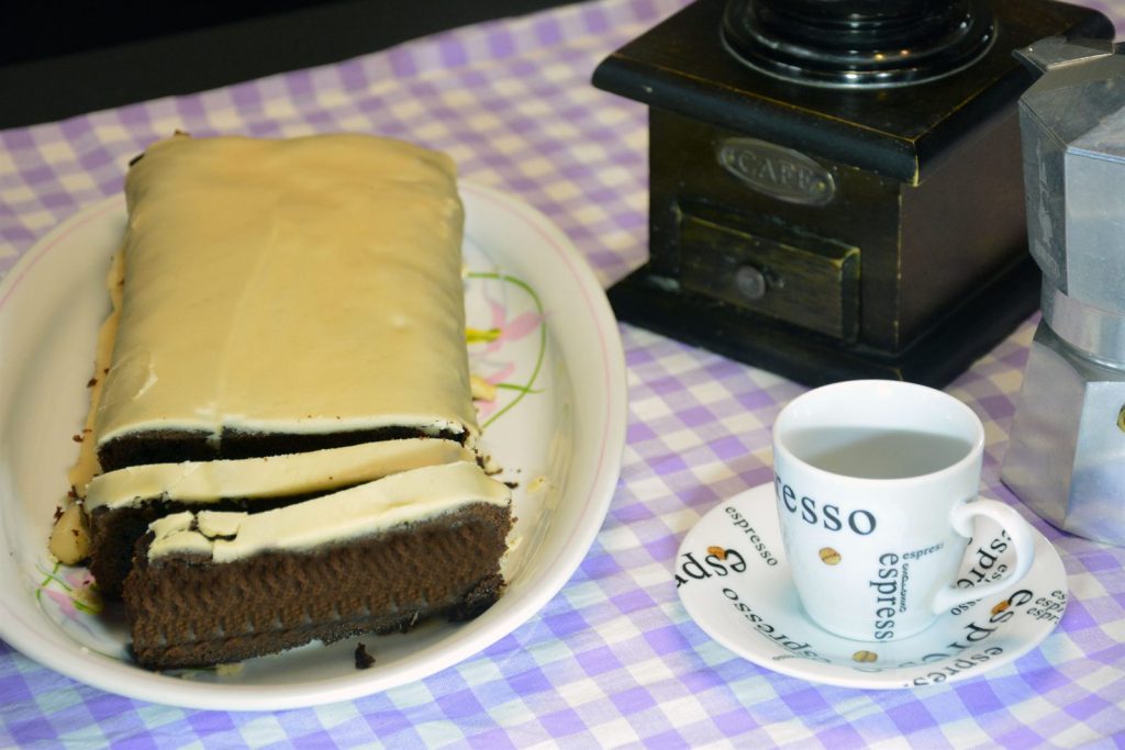 Ciasto kawowe z jasnym lukrem kawowym pokrojone w plastry na białym półmisku, obok filiżanka espresso, stary młynek do kawy i kawiarka