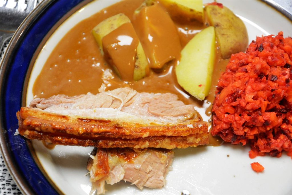 Dwa plastry pieczonego boczku z żeberkami na białym talerzu z ciemnoniebieskim brzegiem; skórka jest złotobrązowa, chrupiąca i warstwowa, a mięso jasne. Za nimi leżą ugotowane ziemniaki w połówkach i ćwiartkach, w skórkach, polane jasnobrązowym sosem, a po prawej kopiec drobno posiekanej surówki w kolorze czerwono-pomarańczowym. Na zdjęciu nie widać sztućców.