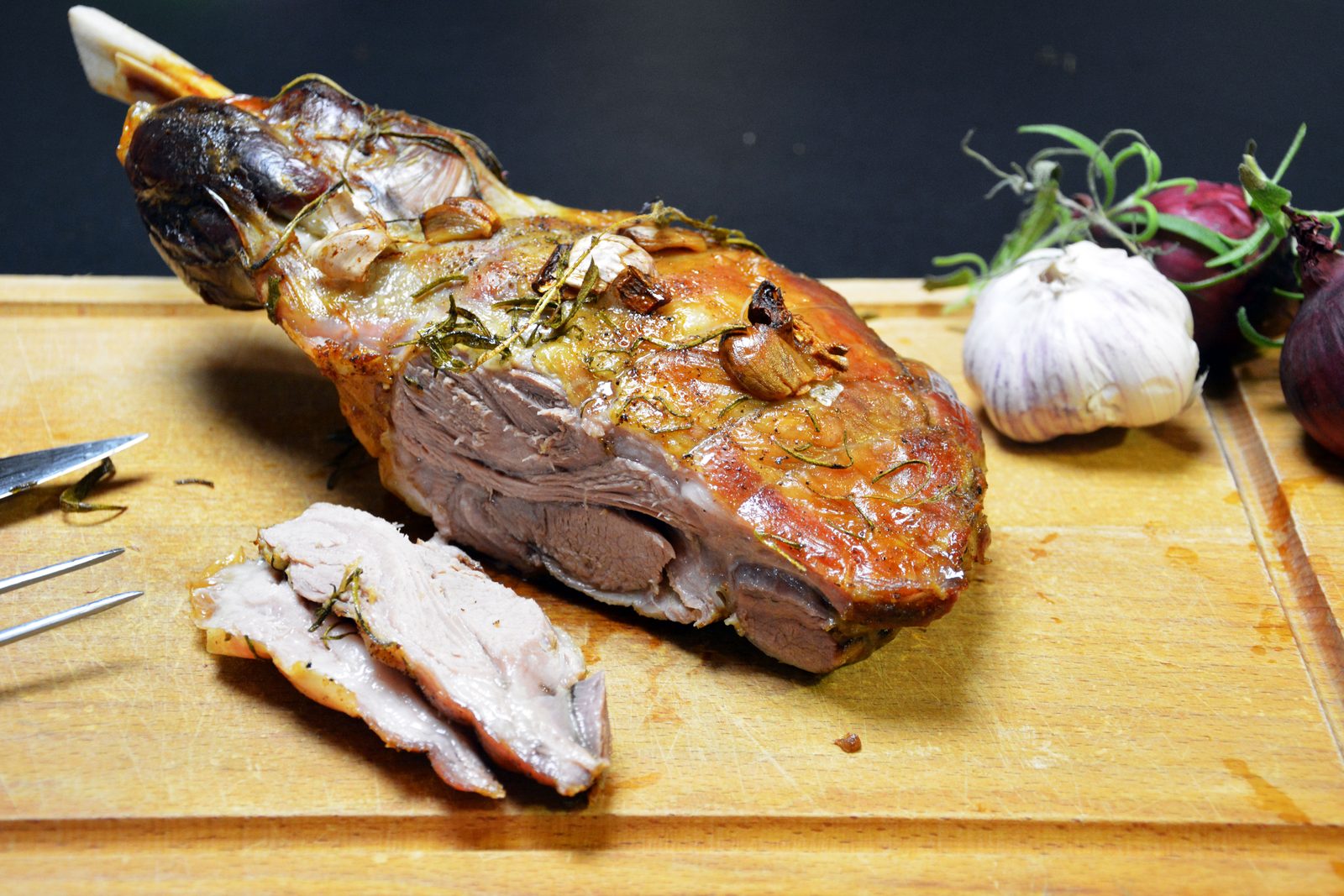 Pieczona łopatka jagnięca z kością, której koniec wystaje po lewej stronie, na drewnianej desce; chrupiąca, brązowa skórka z upieczonymi ząbkami czosnku i igiełkami rozmarynu. Z przodu leżą dwa plastry różowego mięsa, po lewej nóż i widelec, po prawej cała główka czosnku, czerwona cebula i gałązki rozmarynu, ciemne tło.