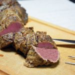 Polędwica wołowa pokrojona w grube plastry leży na jasnej drewnianej desce; mięso jest równomiernie różowe aż po brązową, przypieczoną skórkę. Po prawej stronie leżą nóż do krojenia mięsa i widelec do mięsa z dwoma długimi zębami, w tle białe płótno lub papier, a w prawym górnym rogu ciemne tło.