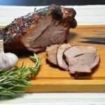 Upieczony na grillu udziec jagnięcy z kością, której koniec wystaje po lewej stronie, na drewnianej desce na pasiastej lnianej serwecie; ciemna, niemal czarna wędzona skórka, przekrojony kawałek odsłania różowe mięso, a przed nim po prawej leżą trzy plastry. Z przodu po lewej cała główka czosnku i gałązki rozmarynu, po prawej nóż i widelec do krojenia.
